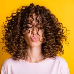 Mujer con un espectacular pelo rizado castaño, haciendo un beso, sobre fondo amarillo vibrante.