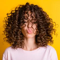 Mujer con pelo rizado fino y escaso, con un peinado voluminoso que cubre su rostro, enviando un beso al aire sobre fondo amarillo.