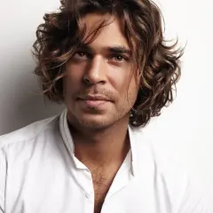 Hombre con peinados media melena, cabello castaño ondulado, mirada intensa y camisa blanca desabrochada.