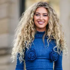 Mujer sonriente con un espectacular pelo chino rubio, luciendo un vestido vaquero azul con detalles de rosas.