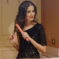 Mujer con vestido negro brillante creando peinados con ondas para bodas, usando una plancha naranja.