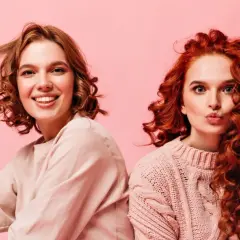 Dos chicas con diferentes tipos de cabello ondulado, una sonríe y juega con su pelo, la otra hace un puchero.