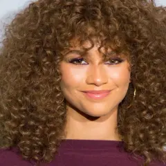 Mujer con cortes pelo rizado largo y voluminoso, maquillaje vibrante y sonrisa radiante.