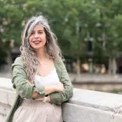 Mujeres pelo rizado con canas, sonriendo con los brazos cruzados, apoyada en un muro de piedra.