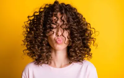 Mujer con un espectacular pelo rizado castaño, haciendo un beso, sobre fondo amarillo vibrante.