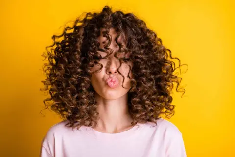 Mujer con un espectacular pelo rizado castaño, haciendo un beso, sobre fondo amarillo vibrante.