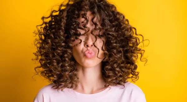Mujer con un espectacular pelo rizado castaño, haciendo un beso, sobre fondo amarillo vibrante.