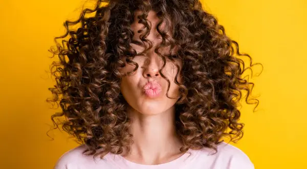 Mujer con pelo rizado fino y escaso, con un peinado voluminoso que cubre su rostro, enviando un beso al aire sobre fondo amarillo.