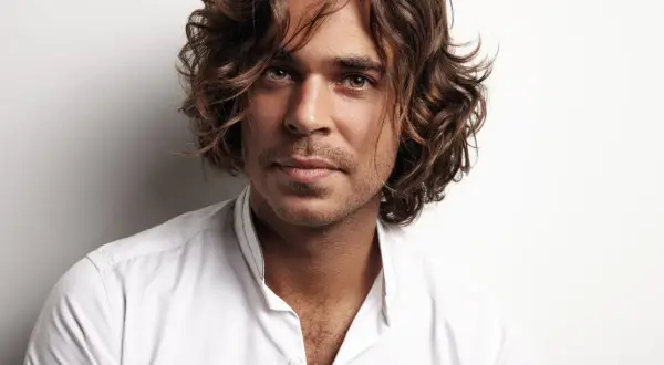 Hombre con peinados media melena, cabello castaño ondulado, mirada intensa y camisa blanca desabrochada.
