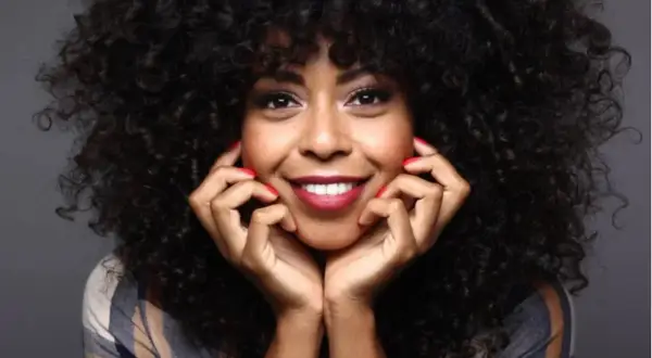 Mujer sonríe con cabello rizado y peinado con finger coils. Sus manos, con uñas rojas, apoyan su rostro.