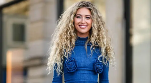 Mujer sonriente con un espectacular pelo chino rubio, luciendo un vestido vaquero azul con detalles de rosas.