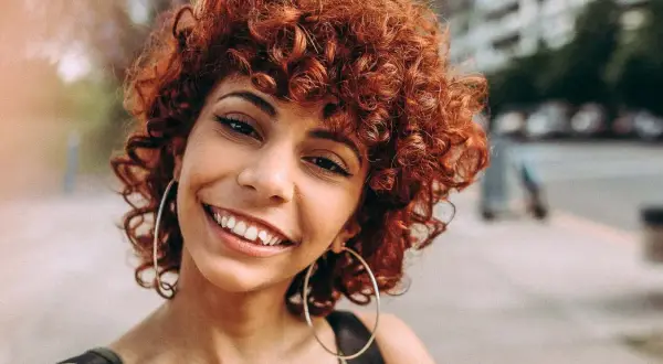 Sonrisa radiante de una mujer con rizos castaño rojizo y grandes aros plateados.