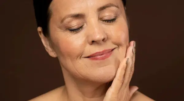 Mujer con ojos cerrados y una mano en la mejilla, sonriendo suavemente. Su piel muestra líneas de expresión, sugiriendo un deseo de quitar arrugas.