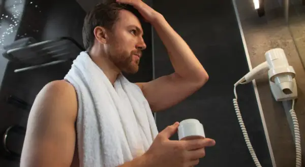 Hombre con pelo fosco se arregla el cabello con crema, envuelto en una toalla blanca.