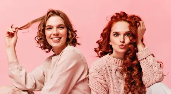 Dos chicas con diferentes tipos de cabello ondulado, una sonríe y juega con su pelo, la otra hace un puchero.
