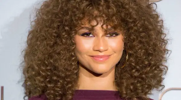 Mujer con cortes pelo rizado largo y voluminoso, maquillaje vibrante y sonrisa radiante.