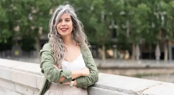 Mujeres pelo rizado con canas, sonriendo con los brazos cruzados, apoyada en un muro de piedra.