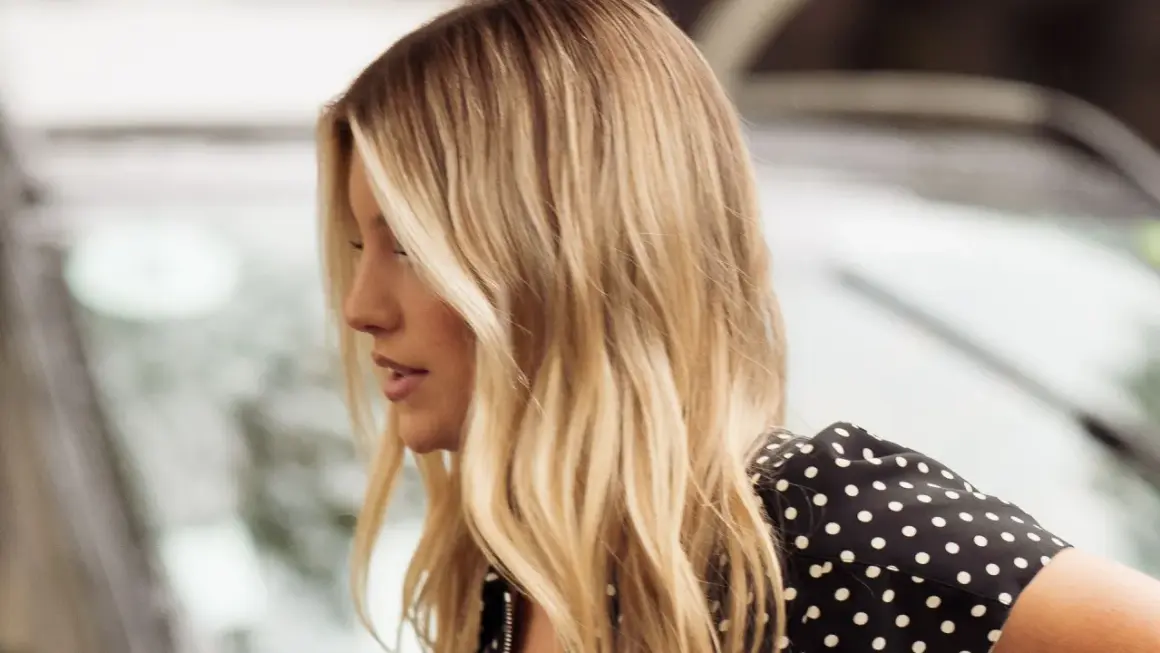 Perfil de mujer con cabello rubio mantequilla, ondas suaves y un vestido negro con lunares blancos.