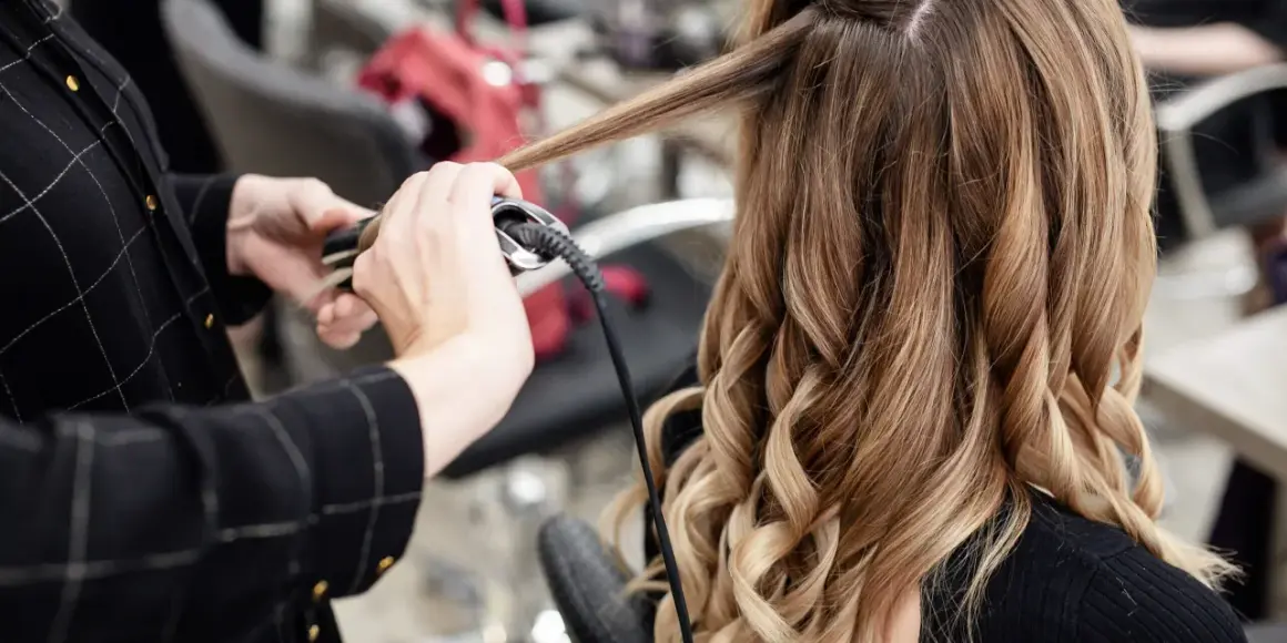 Manos expertas usando una tenaza para rizar el cabello rubio y castaño, creando ondas perfectas.