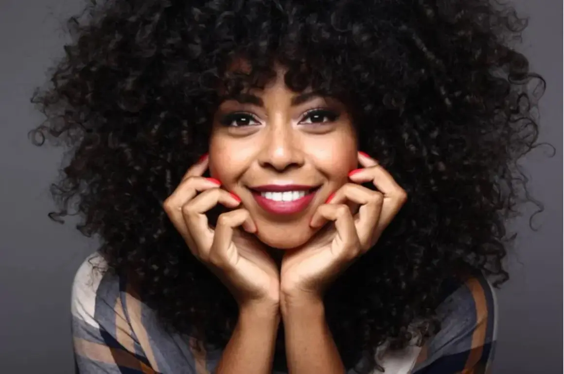 Mujer sonríe con cabello rizado y peinado con finger coils. Sus manos, con uñas rojas, apoyan su rostro.