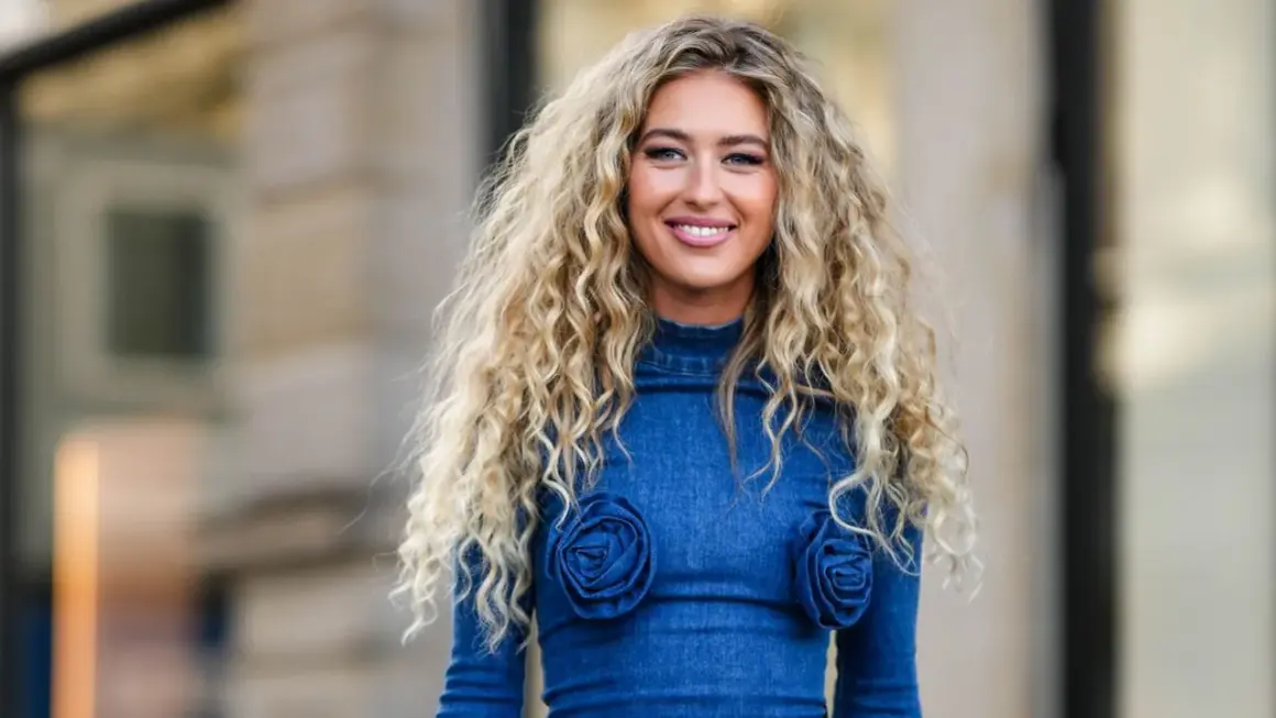 Mujer sonriente con un espectacular pelo chino rubio, luciendo un vestido vaquero azul con detalles de rosas.