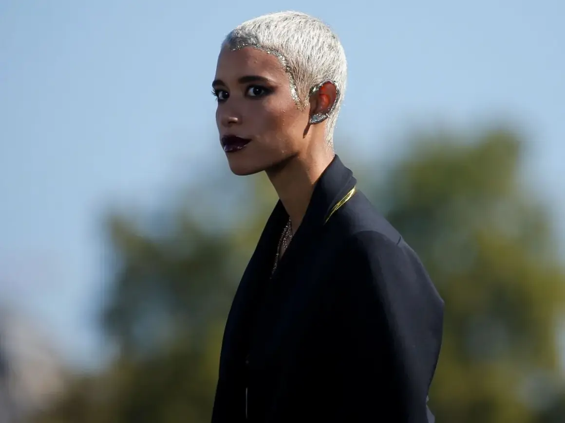 Mujer con pelo blanco corto y maquillaje dramático, luciendo un pendiente llamativo en la oreja.