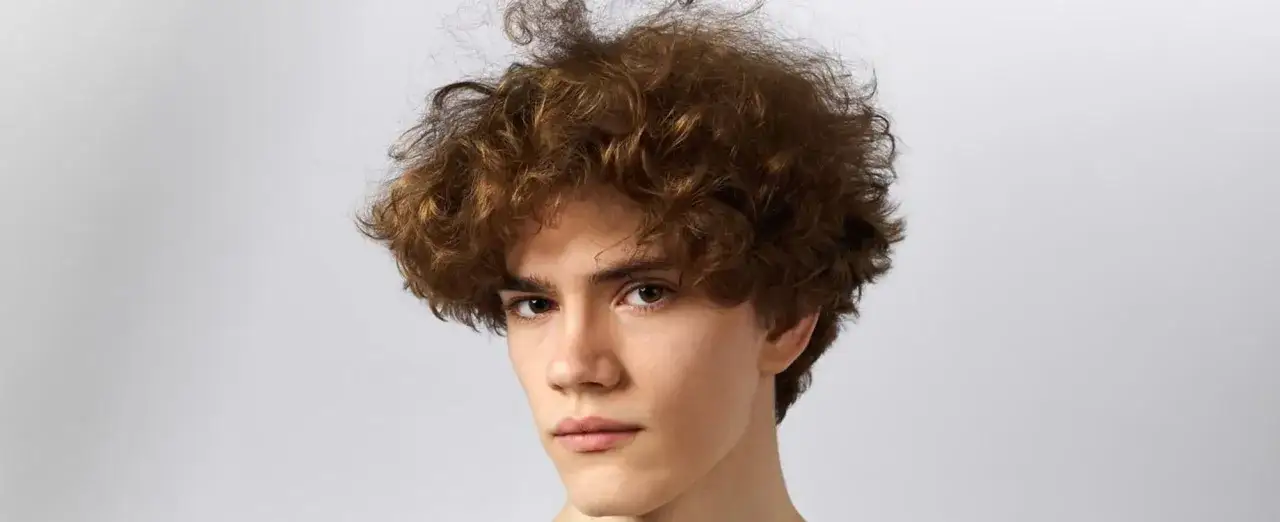Joven con pelo rizado castaño, mirada intensa y fondo neutro.