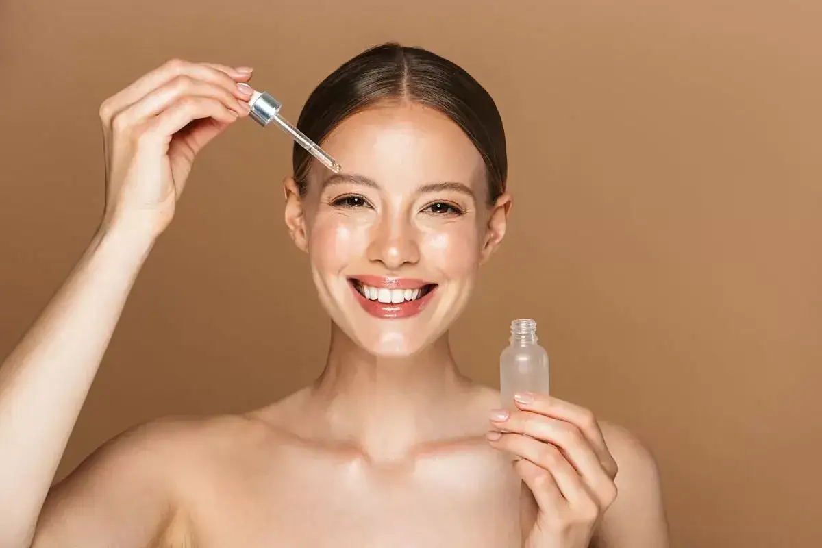 Una mujer sonríe mientras se aplica serum en la frente con un gotero. Muestra cómo se aplica el serum para una piel radiante.