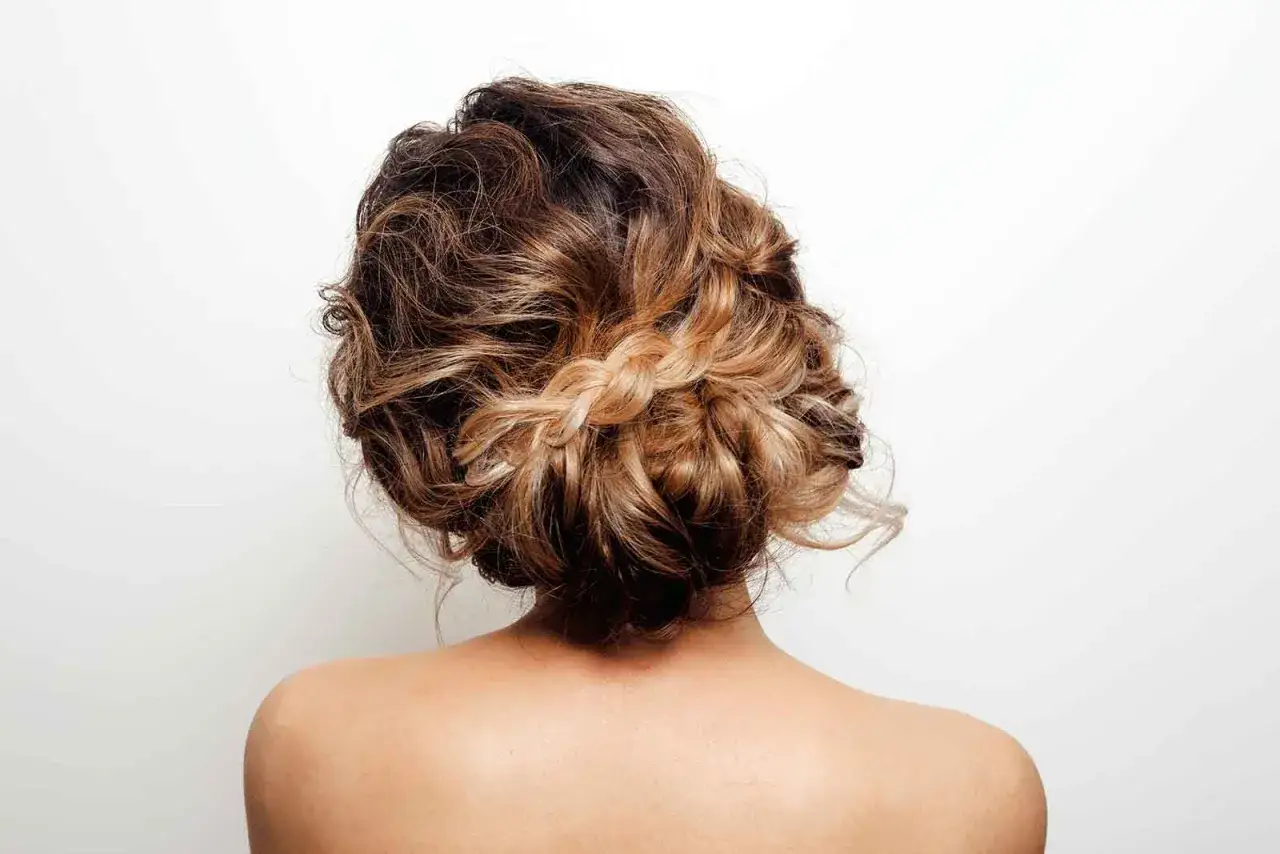 Peinado recogido con trenza en pelo crespo de mujer, con tonos castaños y rubios, sobre fondo blanco.