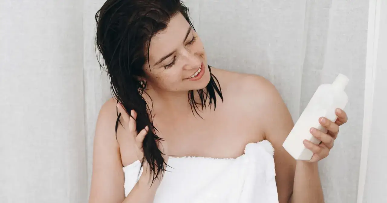 Mujer con cabello mojado y toalla, sosteniendo una botella de acondicionador. Aprende cómo se usa el acondicionador para un cabello suave.