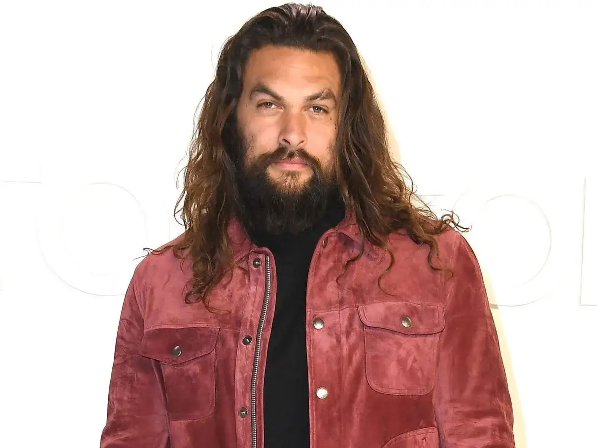 Hombre con pelo largo y barba frondosa, viste una chaqueta de gamuza color vino tinto.
