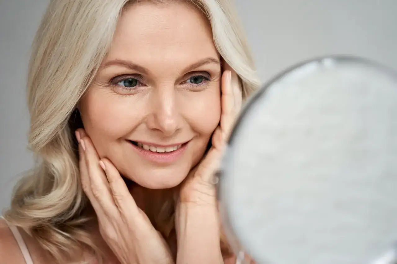 Mujer rubia sonríe, tocando su rostro con las manos. Celebra el pro aging, mostrando confianza y belleza natural.