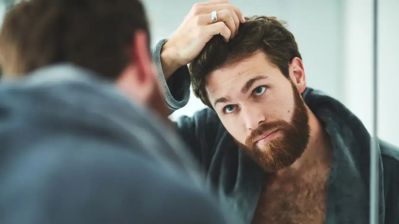 Hombre joven con pelo normal y barba rojiza se mira al espejo, ajustándose el cabello.