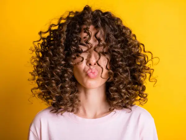 Mujer con pelo rizado fino y escaso, con un peinado voluminoso que cubre su rostro, enviando un beso al aire sobre fondo amarillo.