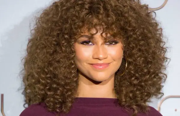 Mujer con cortes pelo rizado largo y voluminoso, maquillaje vibrante y sonrisa radiante.